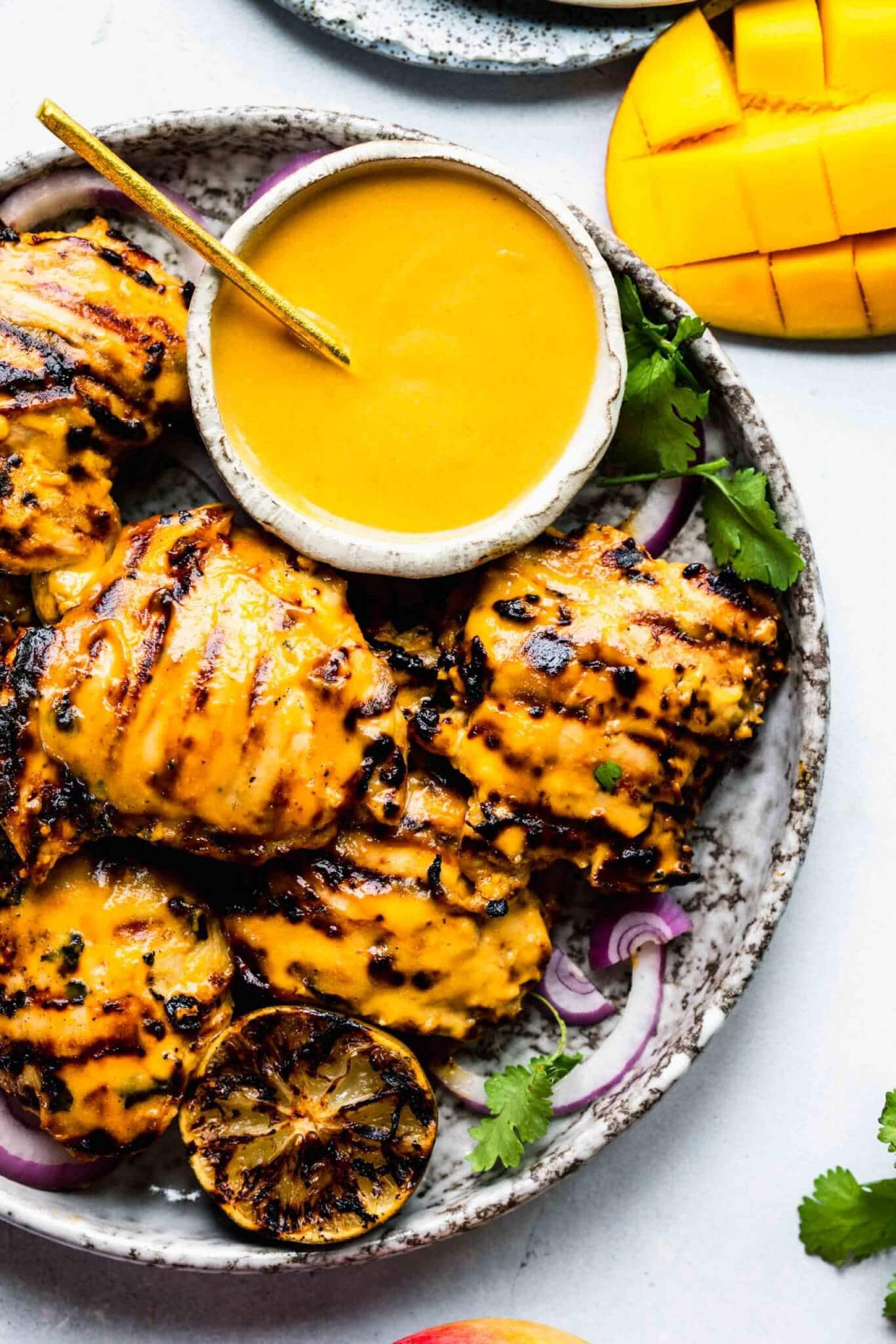 Plate of grilled mango chicken with bowl of mango sauce and charred limes.