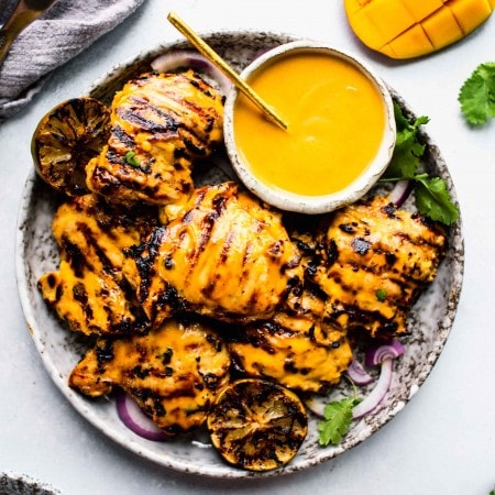 Plate of grilled mango chicken with bowl of mango sauce and charred limes.