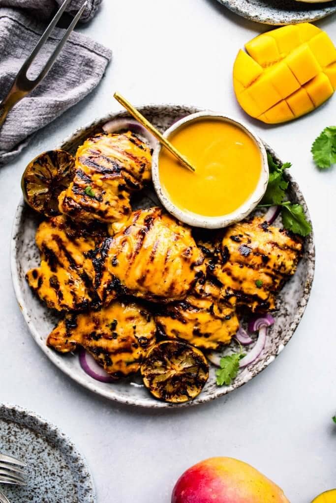 Plate of grilled mango chicken with bowl of mango sauce and charred limes.