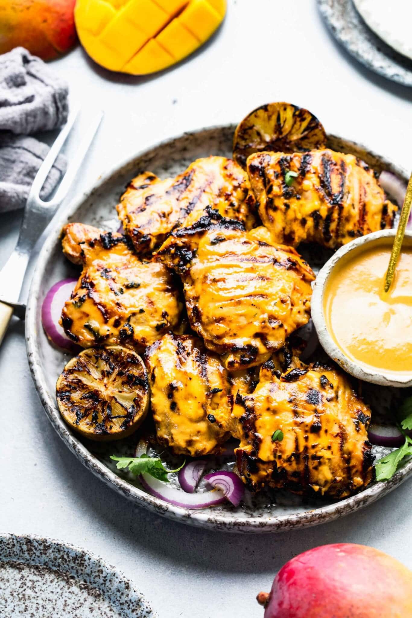 Side view of Plate of grilled mango chicken with bowl of mango sauce and charred limes.