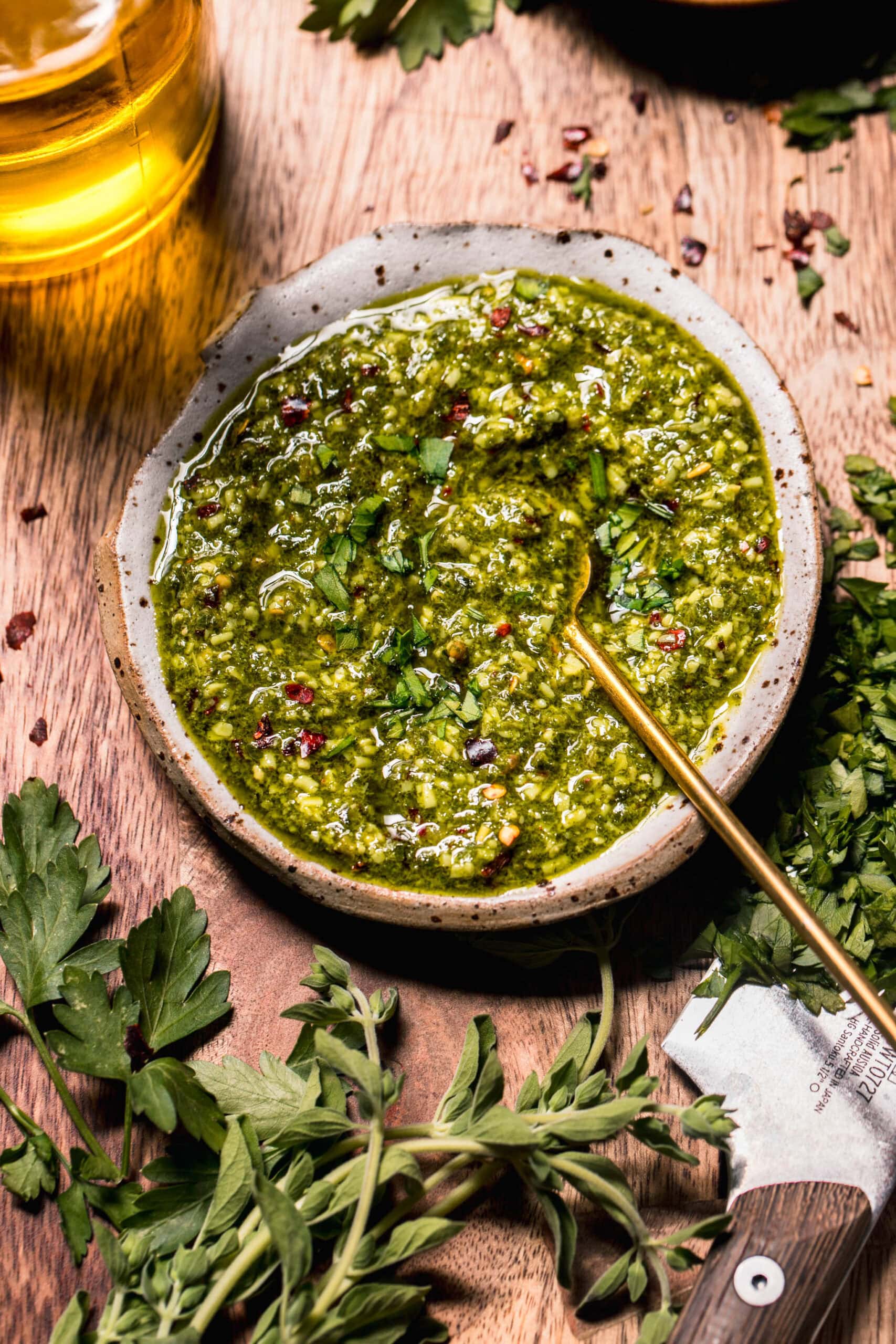 Overhead shot of chimichurri sauce in small rustic bowl on wood cutting board with fresh herbs.