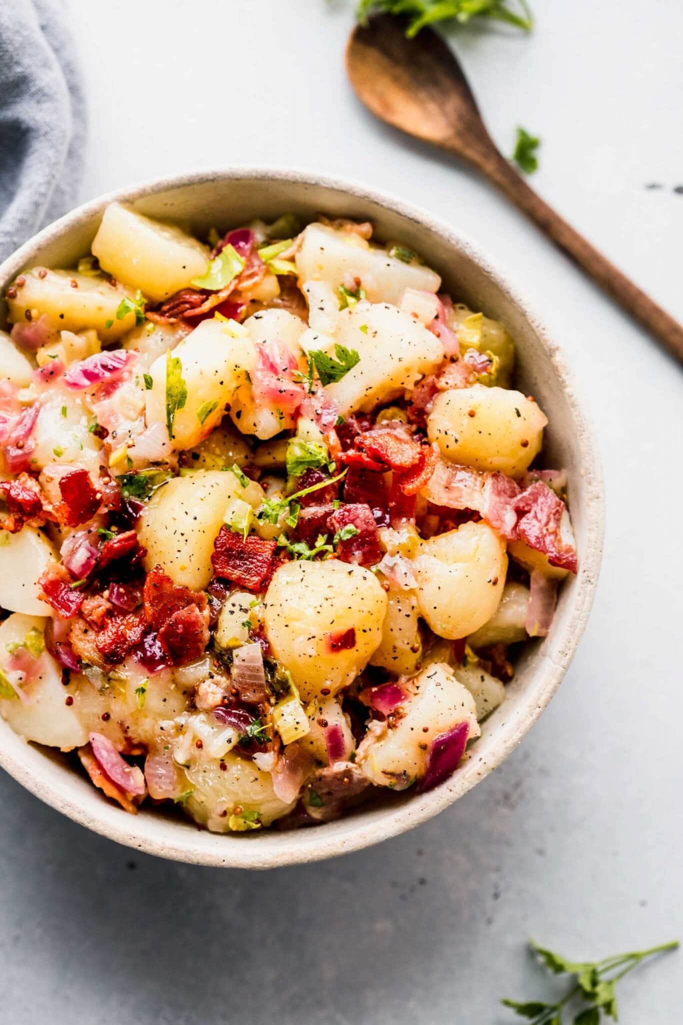 German potato salad in white bowl next to wooden spoon.