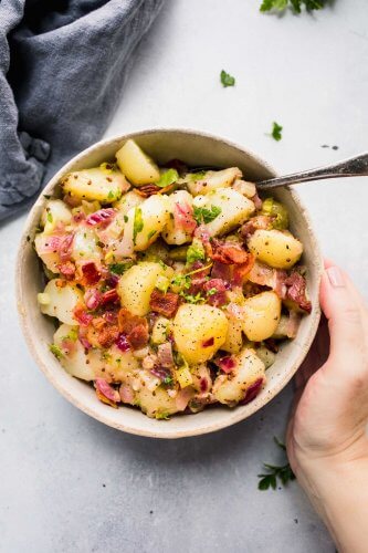 Hand holding German potato salad in white bowl next to wooden spoon.