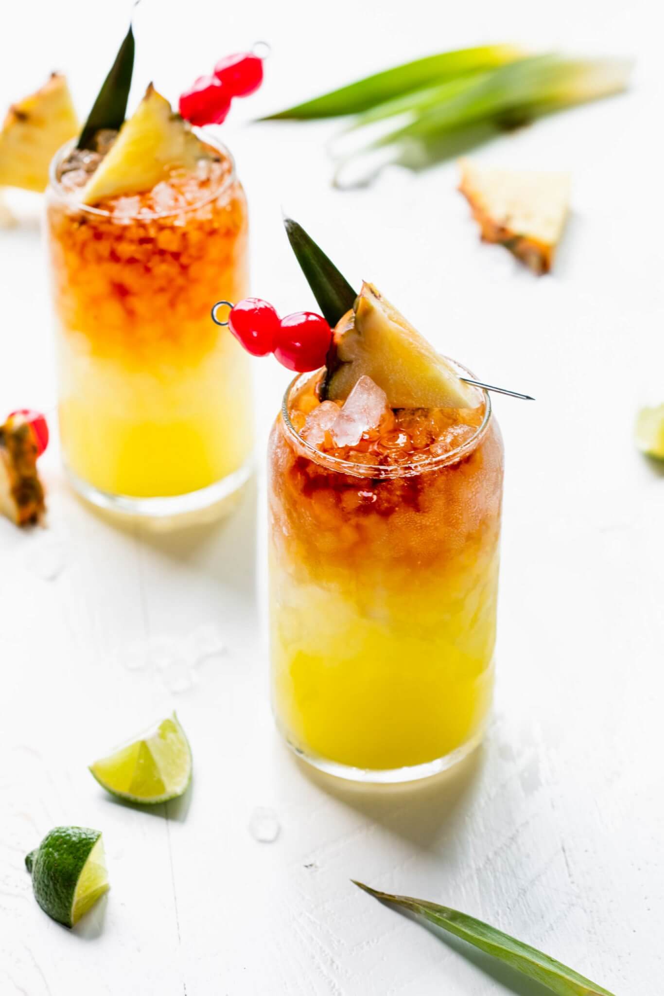 Two Mai tai cocktails in glass garnished with pineapple wedge and cherries.