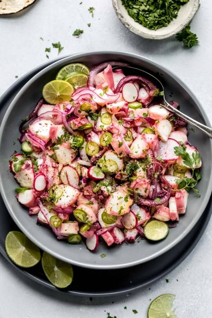 Overhead shot of octopus ceviche in large grey bowl next to lime slices and tortillas.