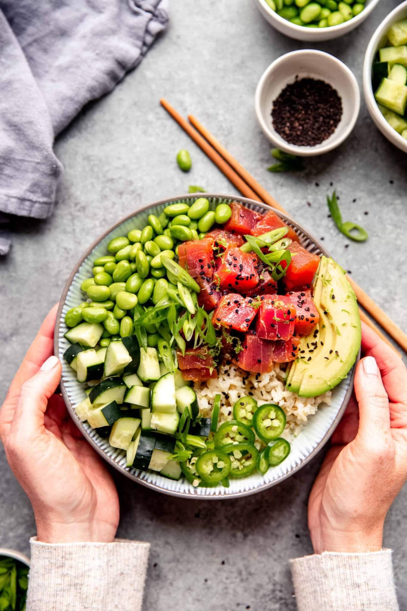 Two hands holding poke bowl next to small bowl of soy sauce and chopsticks.
