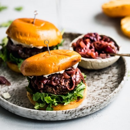 Side view of two lamb burgers arranged on plate next to bowl of caramelized onions and glass of red wine.