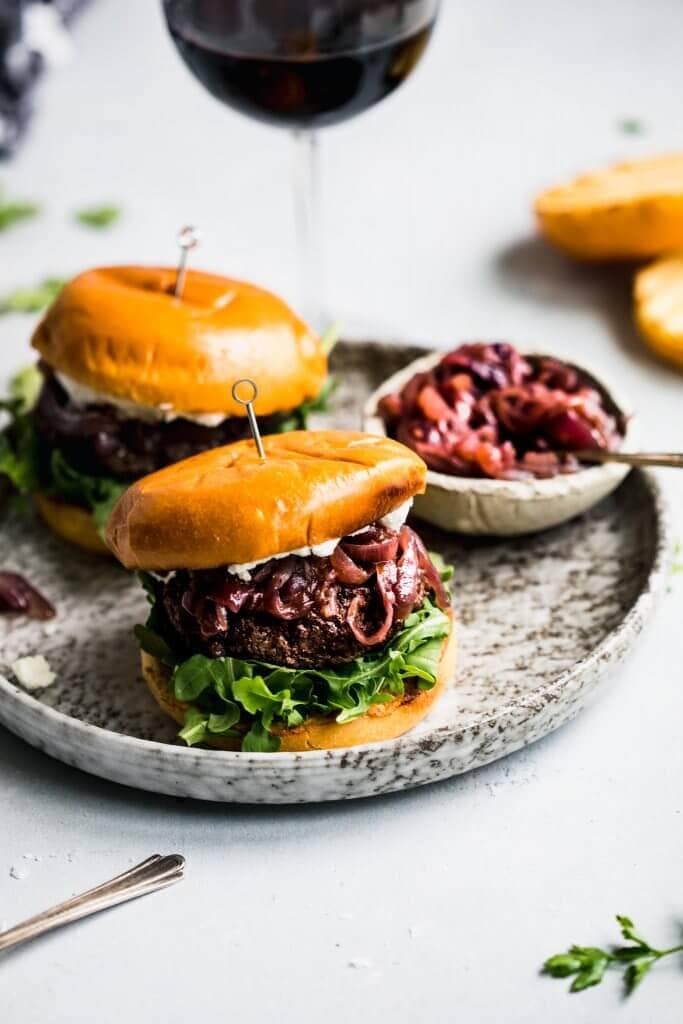 Side view of two lamb burgers arranged on plate next to bowl of caramelized onions and glass of red wine.