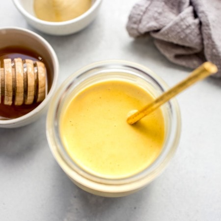 Overhead shot of small glass jar of honey mustard sauce next to honey wand and small bowl of mustard