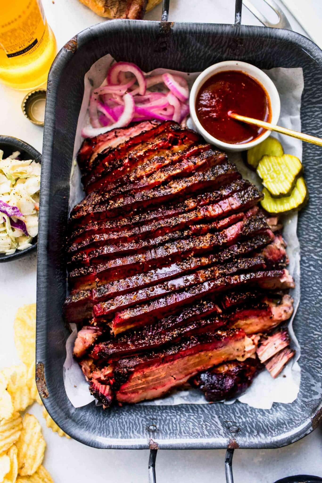 Overhead shot of sous vide cooked brisket on serving tray next to pickles, bbq sauce and buns.