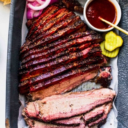 Overhead shot of sous vide cooked brisket on serving tray next to pickles, bbq sauce and buns.