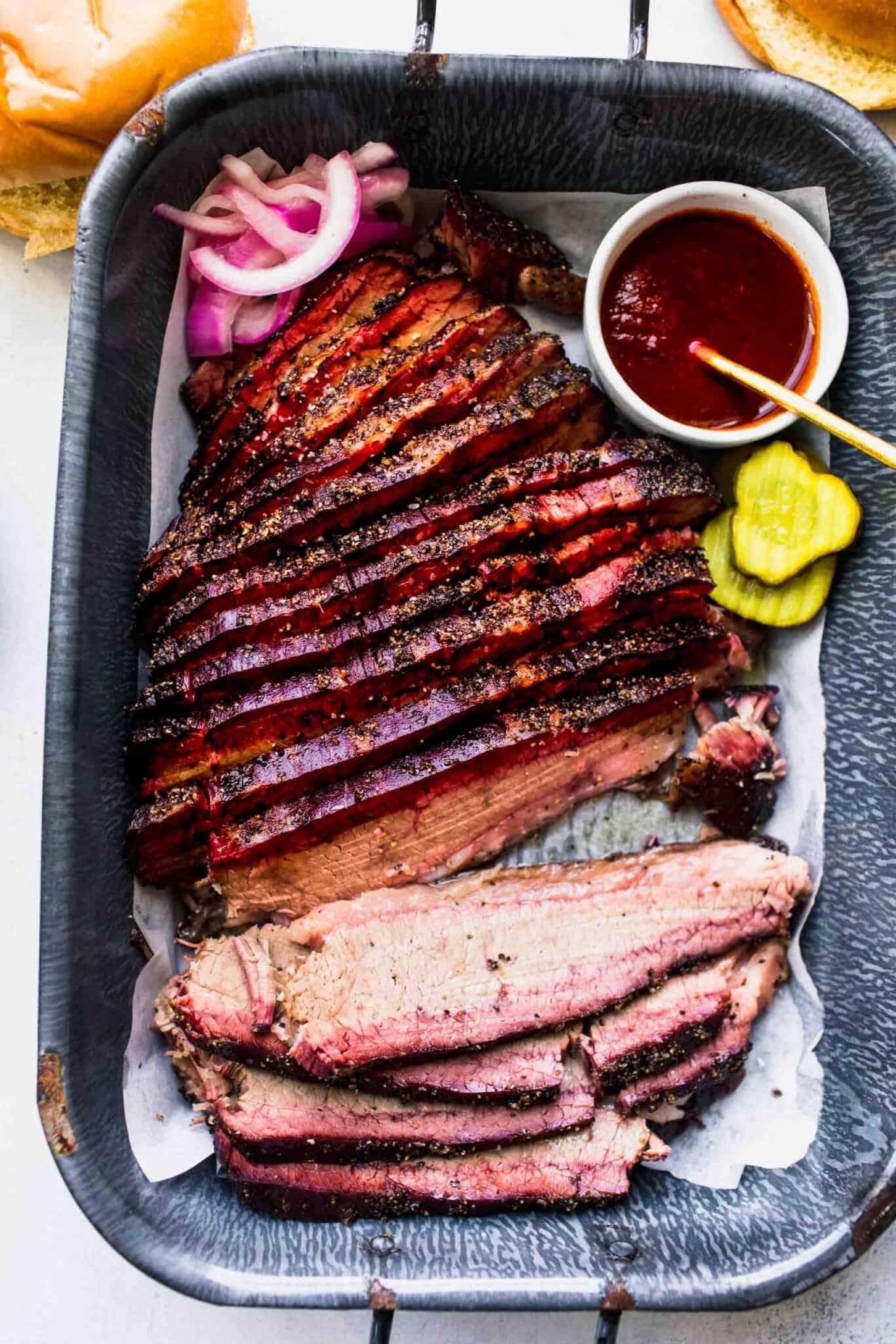 Overhead shot of sous vide cooked brisket on serving tray next to pickles, bbq sauce and buns.