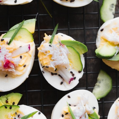 Stuffed crab deviled eggs on dark background topped with sesame seeds, avocado, radishes and cucumber.