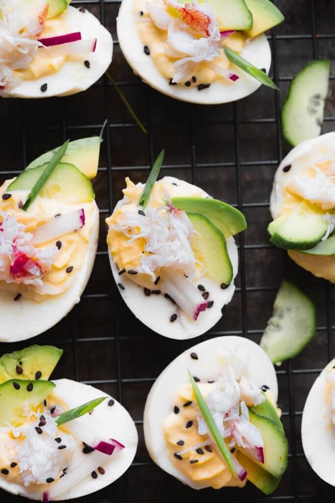 Stuffed crab deviled eggs on dark background topped with sesame seeds, avocado, radishes and cucumber.