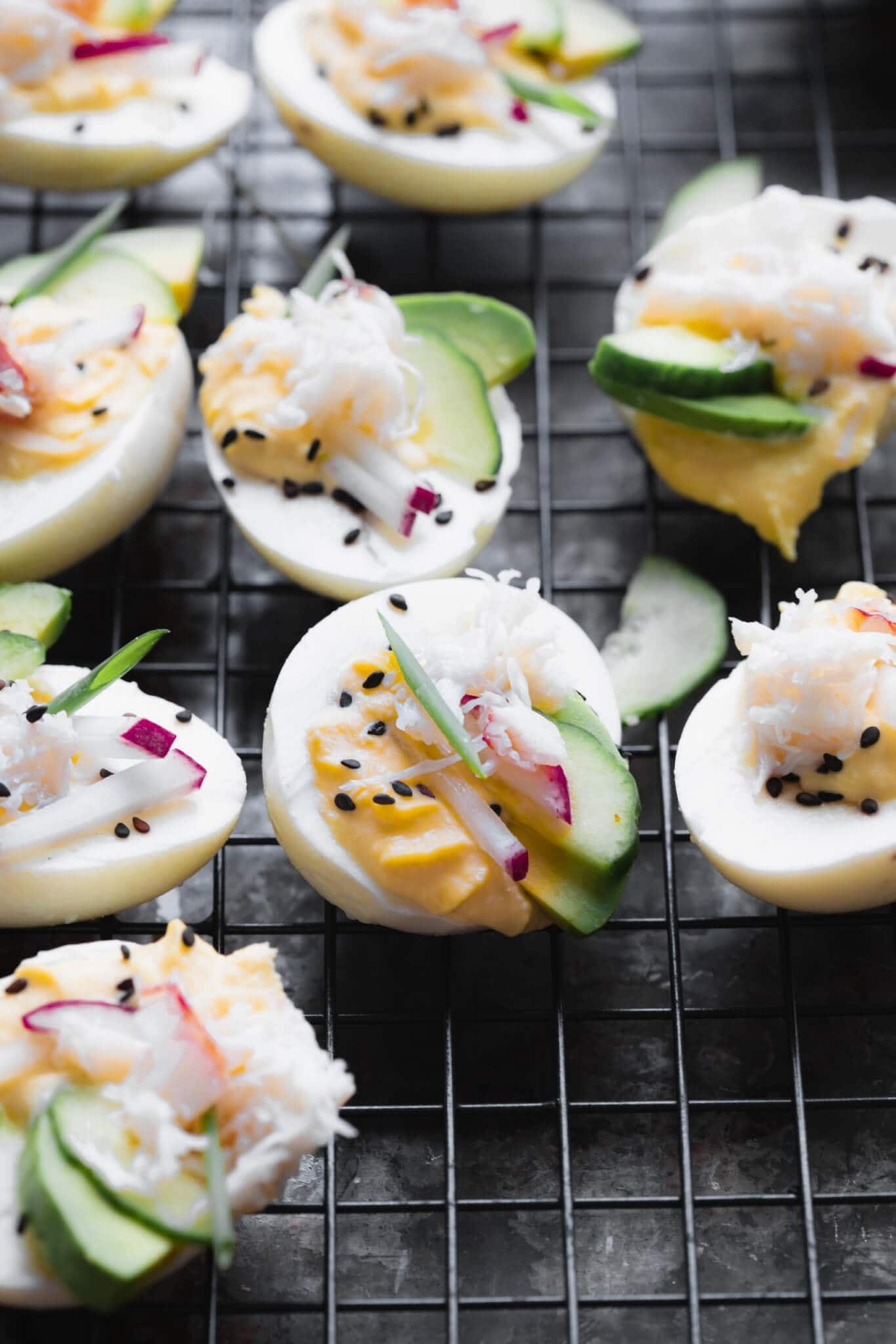 Side view of stuffed crab deviled eggs on dark background topped with sesame seeds, avocado, radishes and cucumber. 