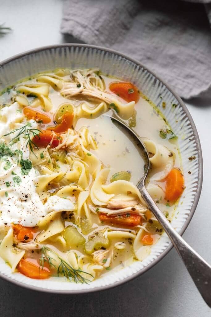 Chicken noodle soup in bowl with spoon, topped with sour cream, dill and parsley.