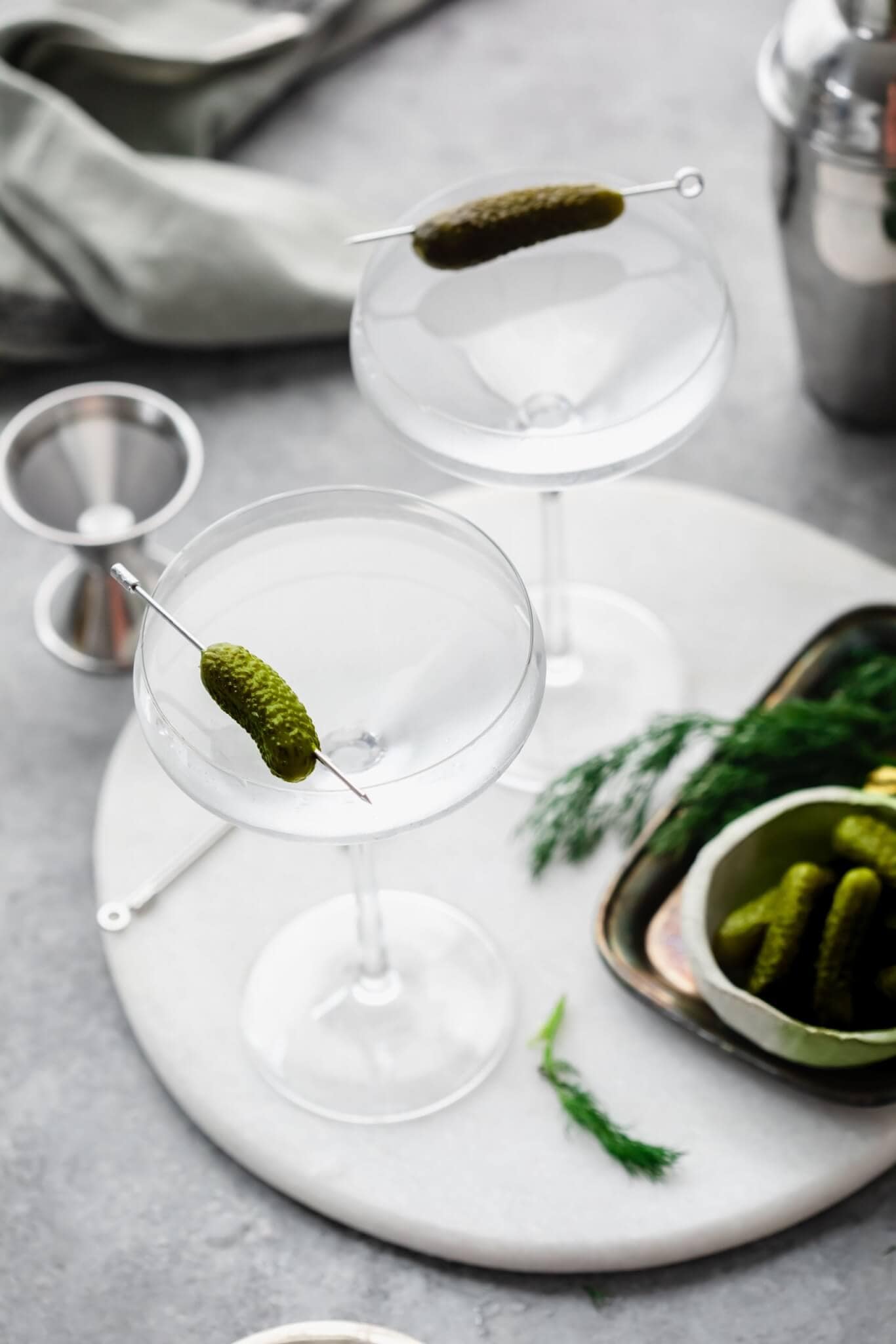 Two pickle martinis on white tray next to shot glass and small bowl of pickles. 