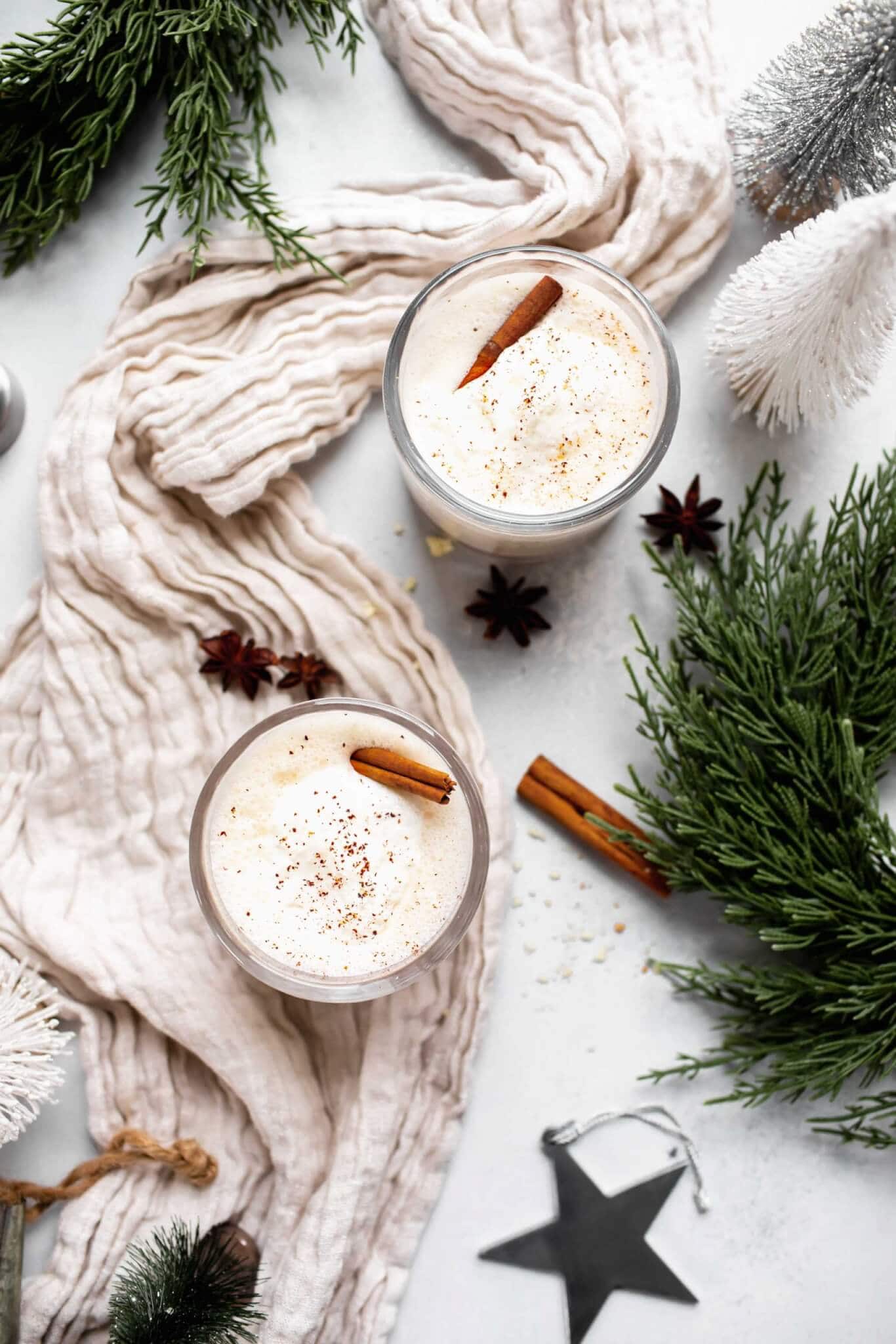Two glasses of eggnog topped with whipped cream and sprinkle of nutmeg. Garnished with cinnamon stick. On table with christmas ornaments and greenery.