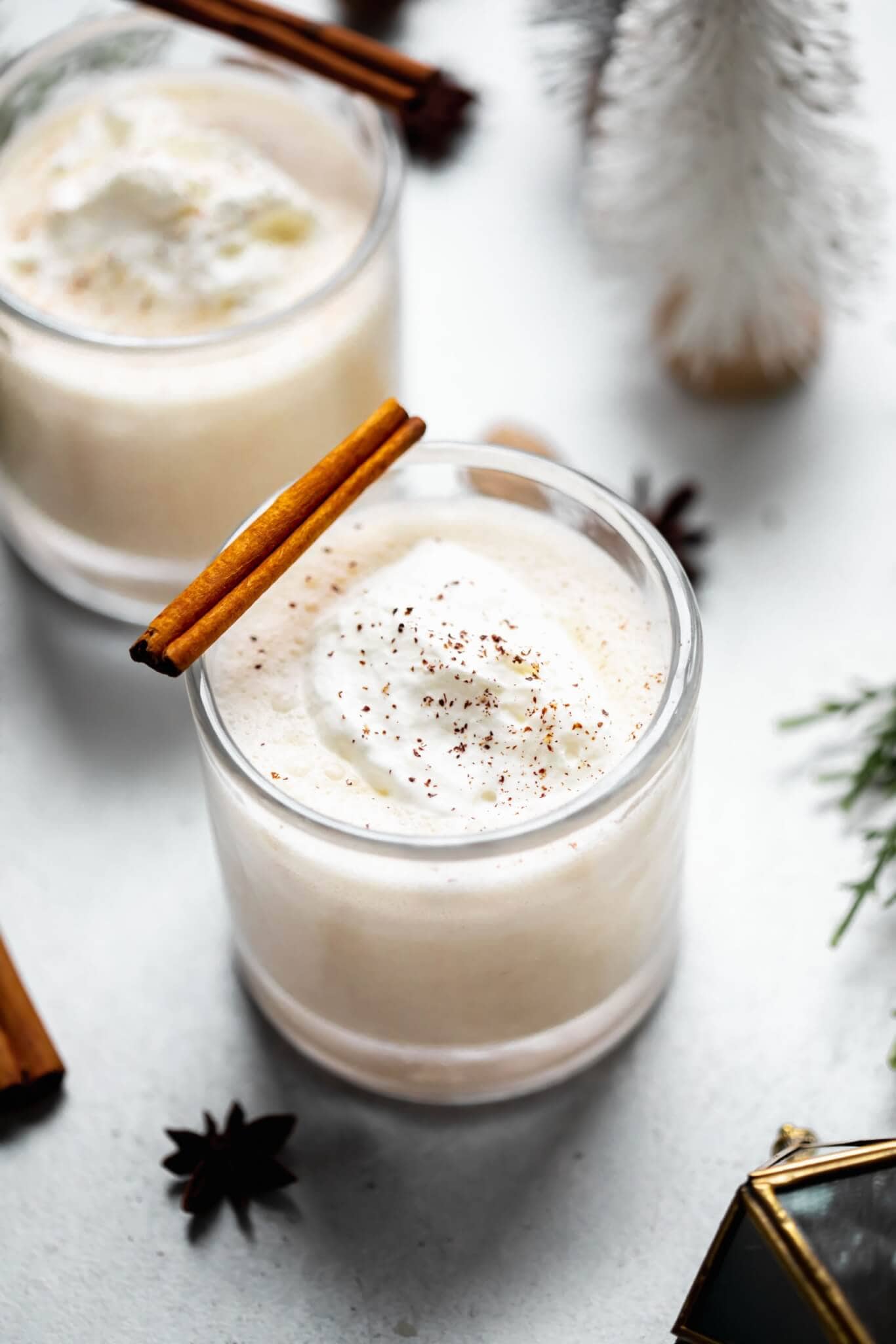 Two glasses of eggnog topped with whipped cream and sprinkle of nutmeg. Garnished with cinnamon stick.