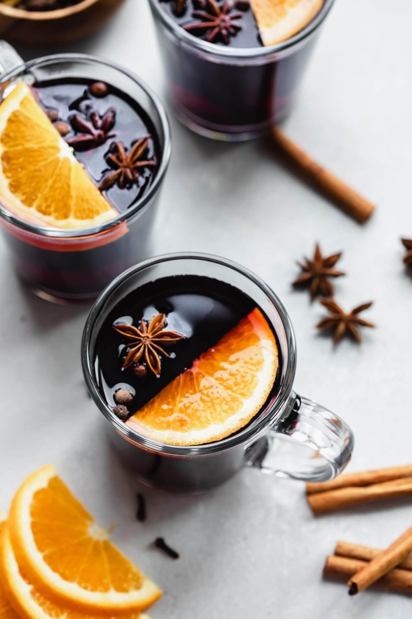 Three mugs of mulled wine on counter next to cinnamon sticks and orange slices.