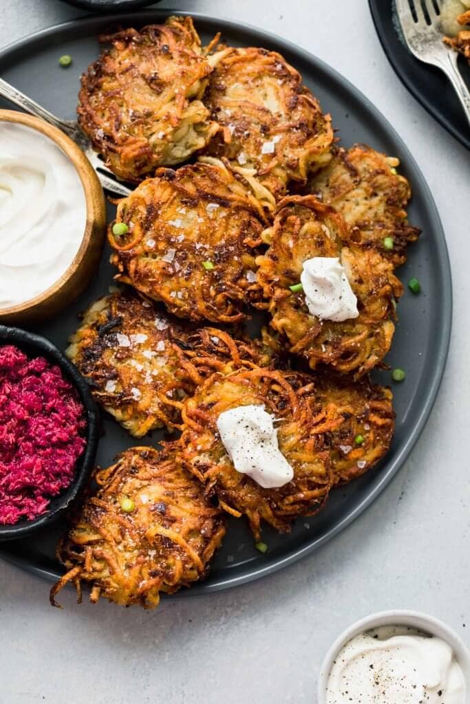 POTATO LATKES ON GREY PLATE TOPPED WITH DOLLOPS OF SOUR CREAM AND SERVED WITH BOWL OF PINK BEET HORSERADISH SAUCE.