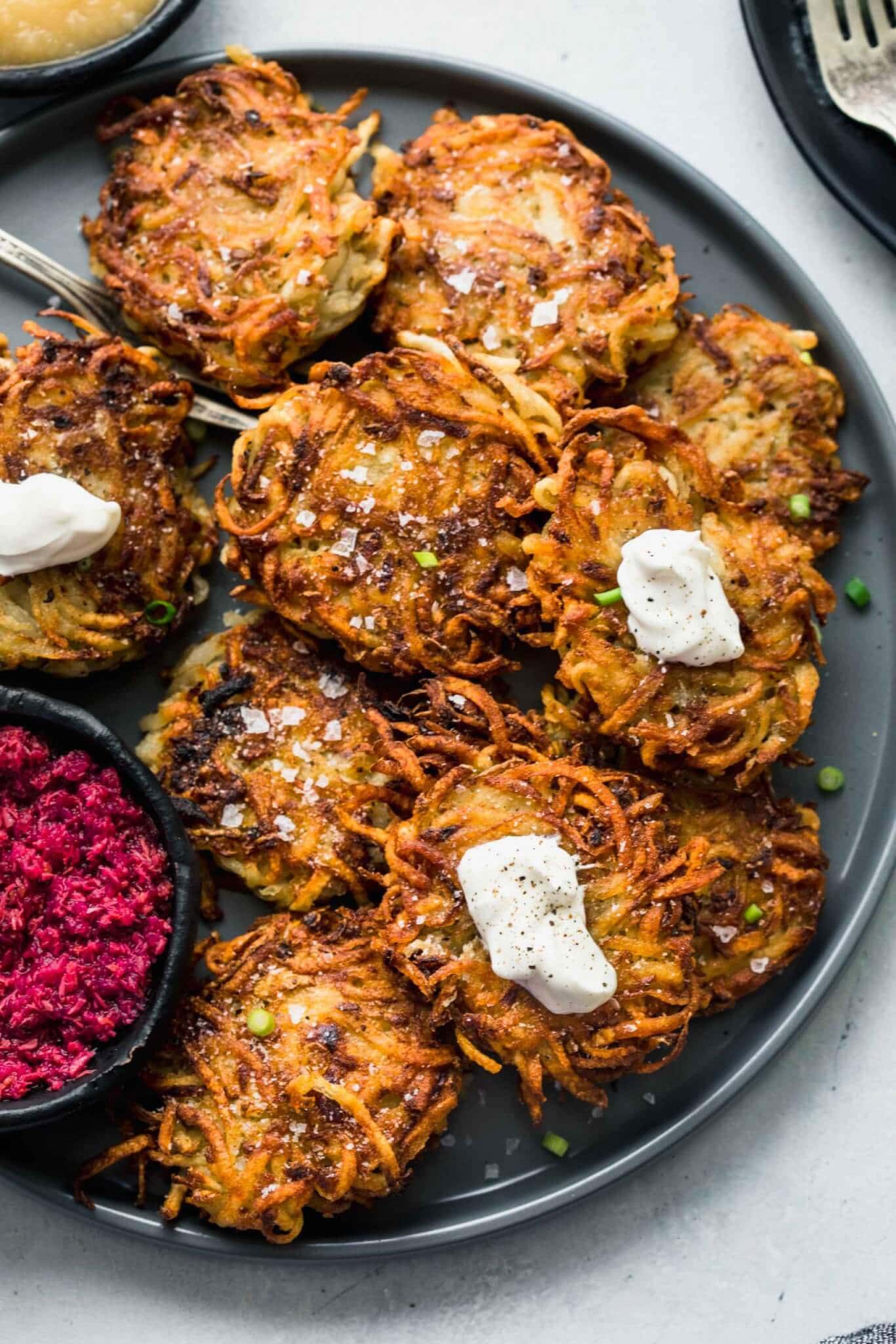 POTATO LATKES ON GREY PLATE TOPPED WITH DOLLOPS OF SOUR CREAM AND SERVED WITH BOWL OF PINK BEET HORSERADISH SAUCE.