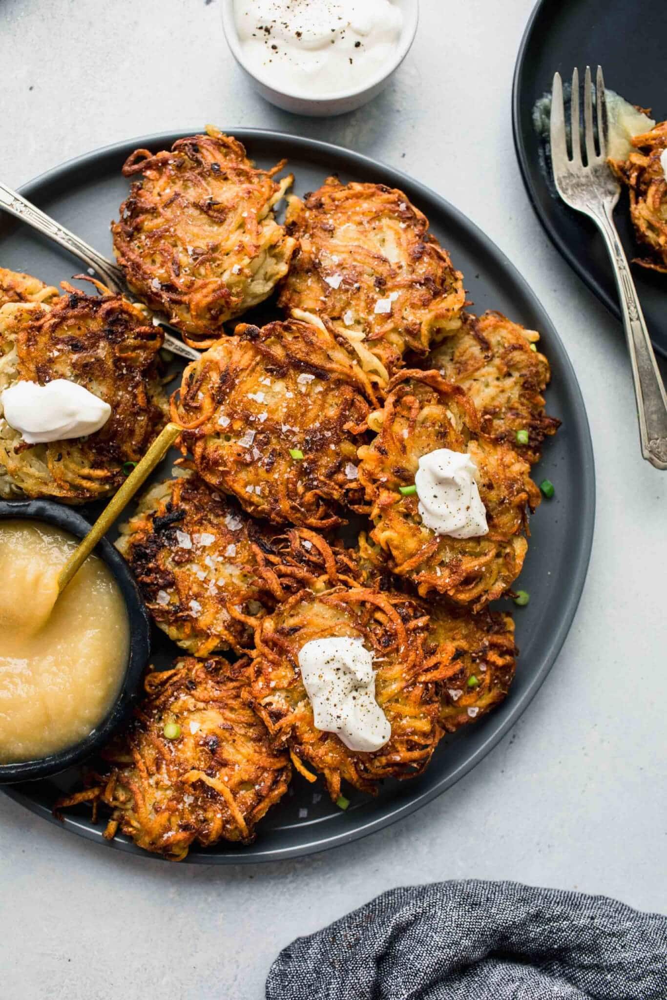 POTATO LATKES ON GREY PLATE NEXT TO SMALL BOWL OF APPLESAUCE AND SOUR CREAM.