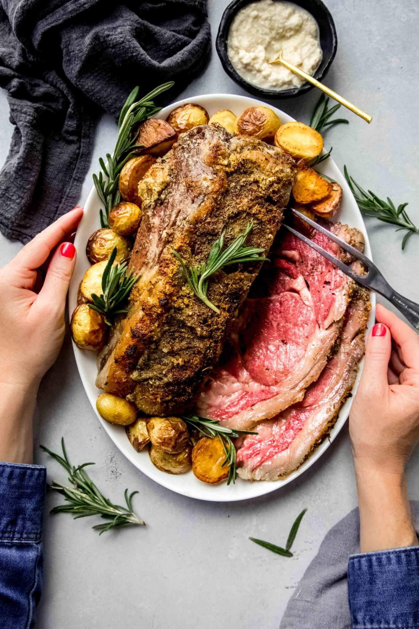 Two hands holding platter or prime rib arranged with roasted potatoes.