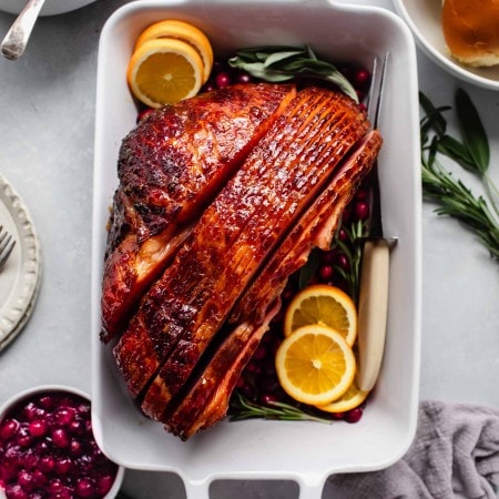 Glazed ham in roasting dish garnished with orange slices, served with mashed potatoes, cranberry sauce and rolls.