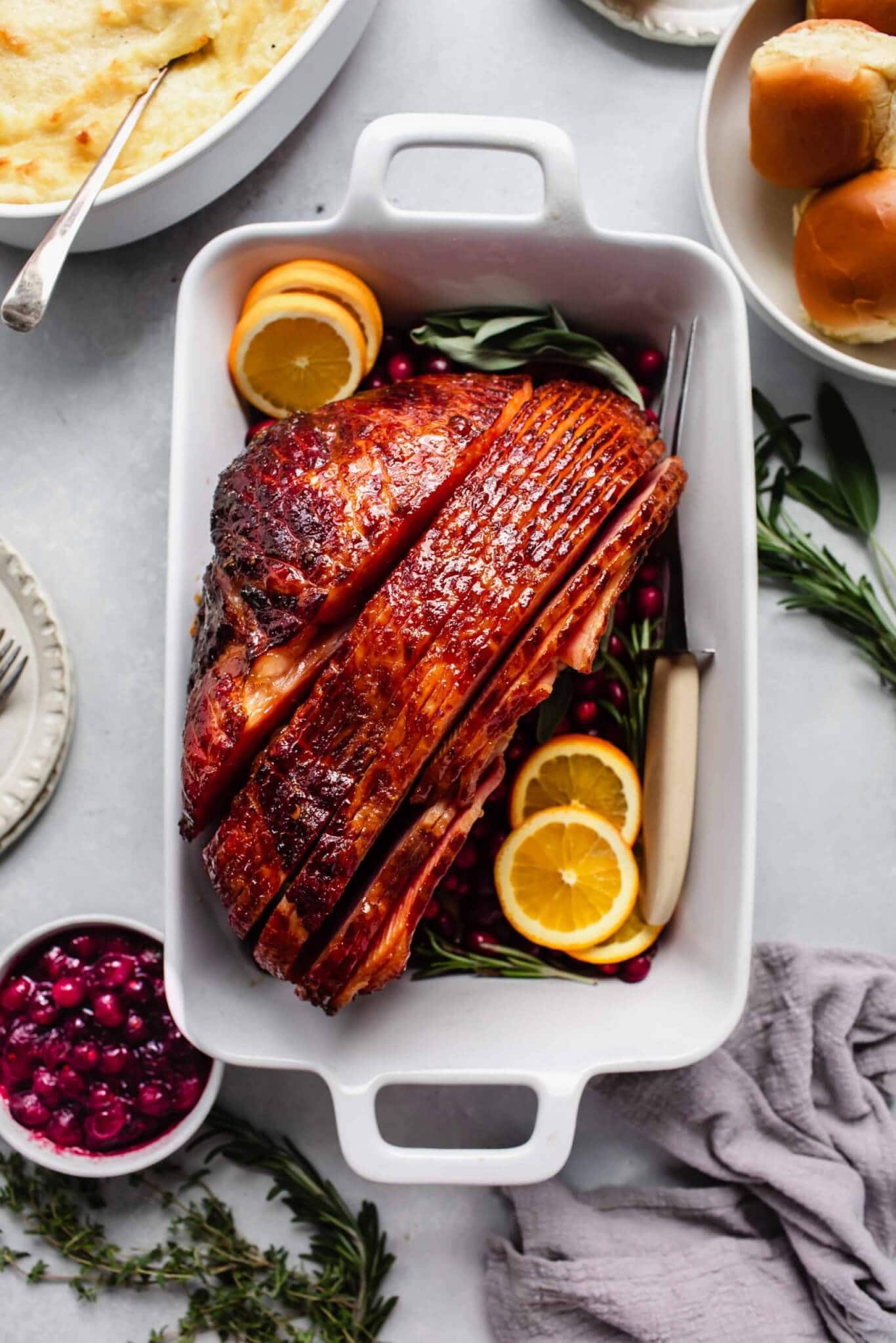 Glazed ham in roasting dish garnished with orange slices, served with mashed potatoes, cranberry sauce and rolls.