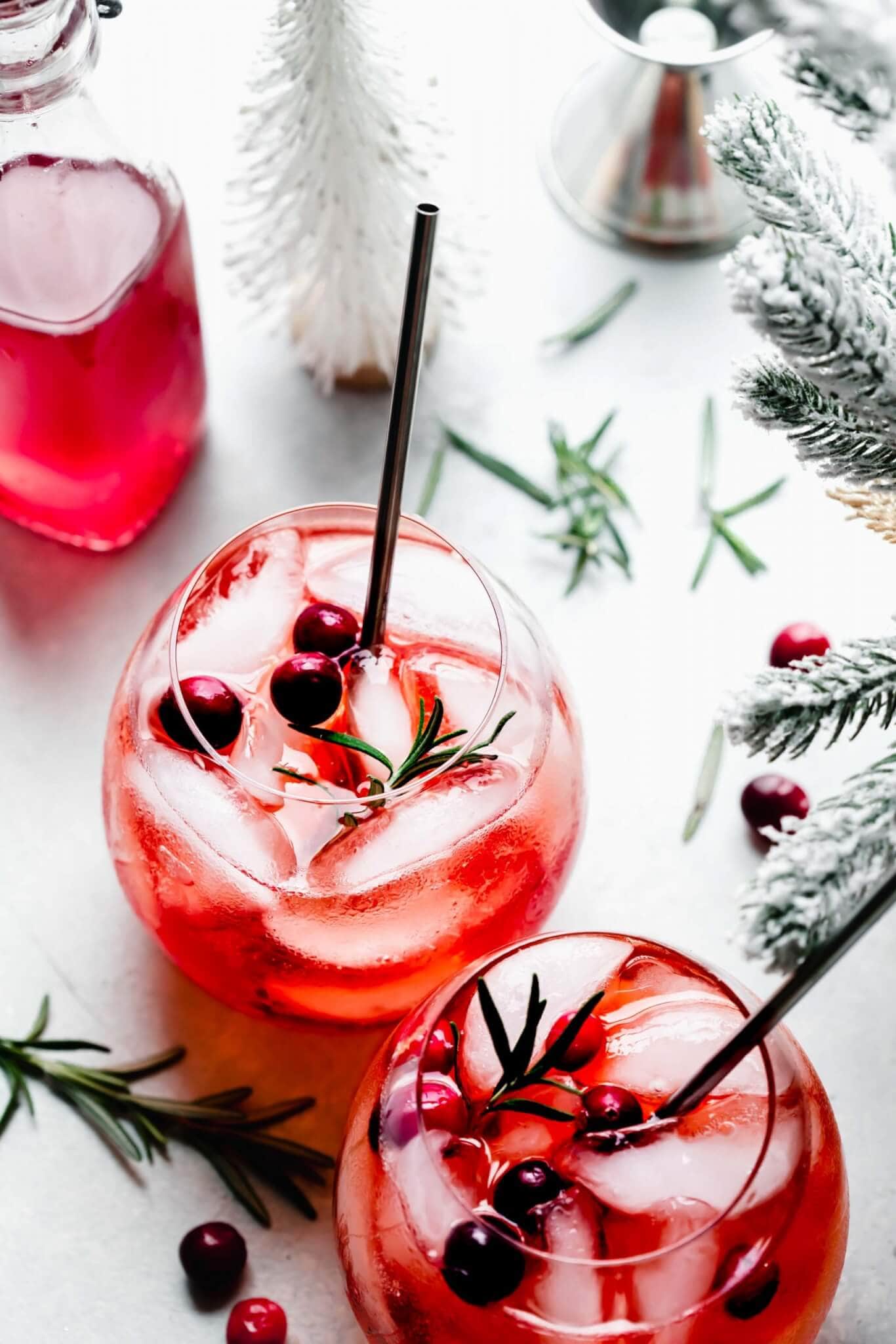 Two winter aperol spritz cocktails next to flocked christmas tree and bottle of cranberry simple syrup.