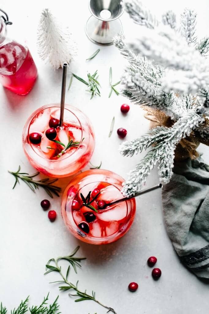 Two winter aperol spritz cocktails next to flocked christmas tree and bottle of cranberry simple syrup.