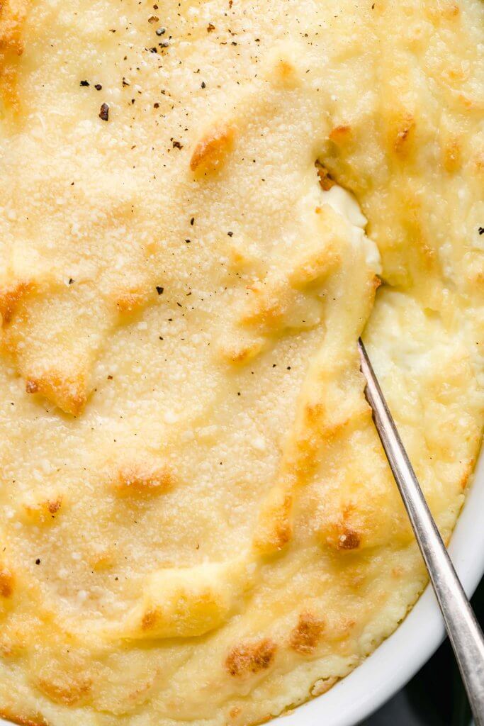 Overhead shot of baked goat cheese mashed potatoes in white casserole dish with serving spoon.