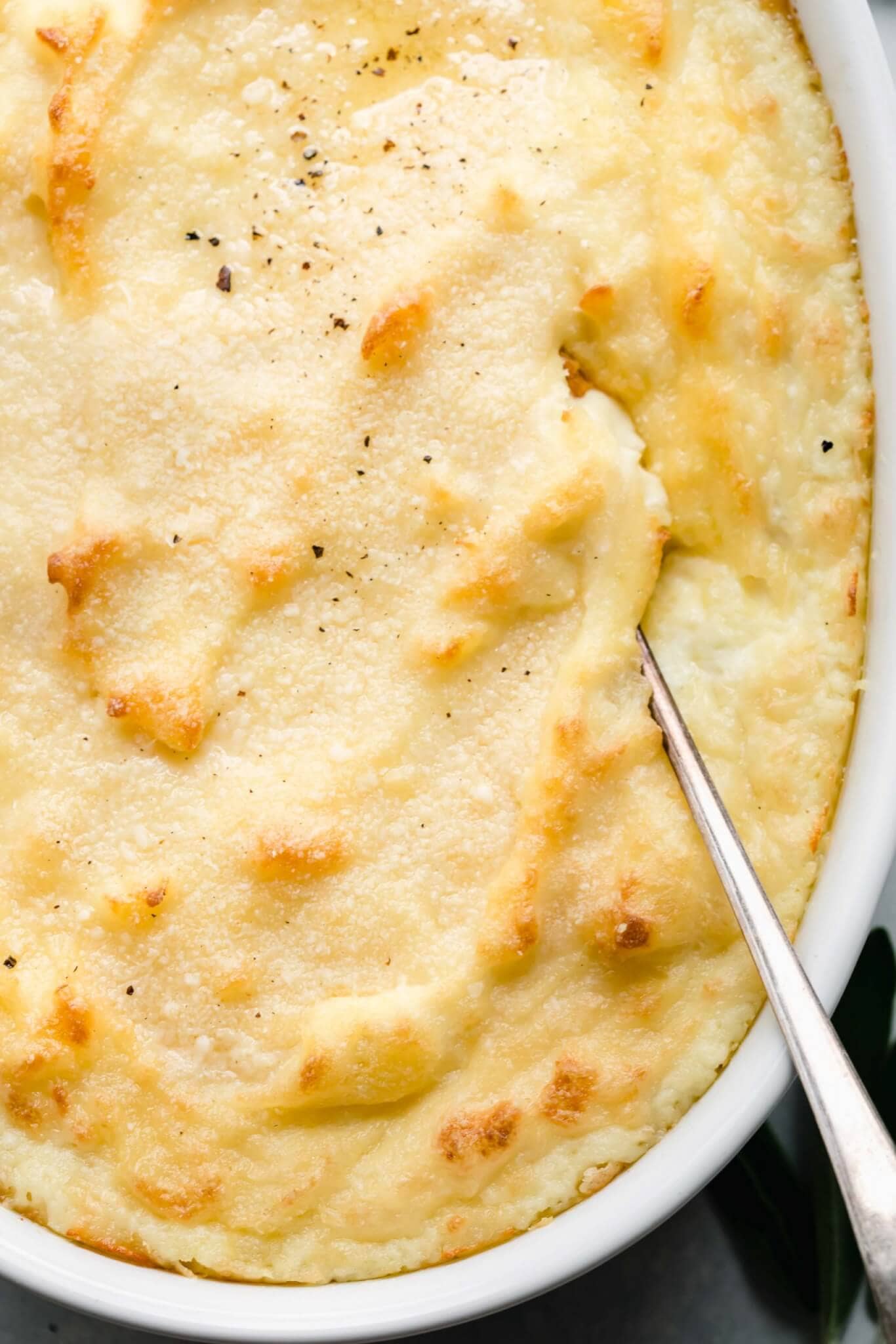 Overhead shot of baked goat cheese mashed potatoes in white casserole dish with serving spoon.