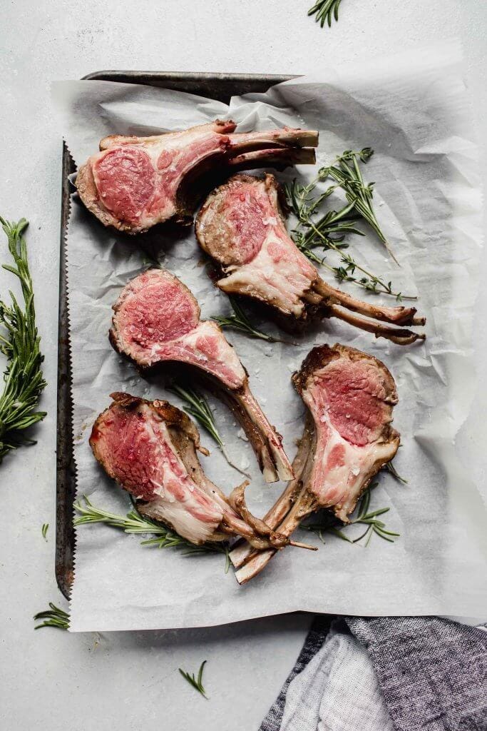 Rack of lamb sliced into chops and laid out on tray next to fresh herbs.