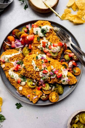 Tortilla chip crusted chicken cutlets arranged on plate topped with queso, tomatoes and red onion.