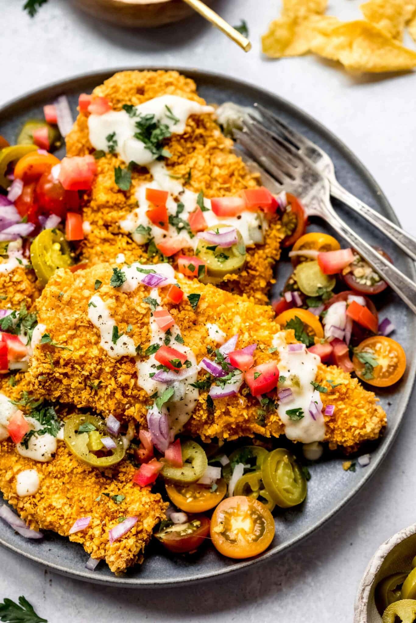 Tortilla chip crusted chicken cutlets arranged on plate topped with queso, tomatoes and red onion.