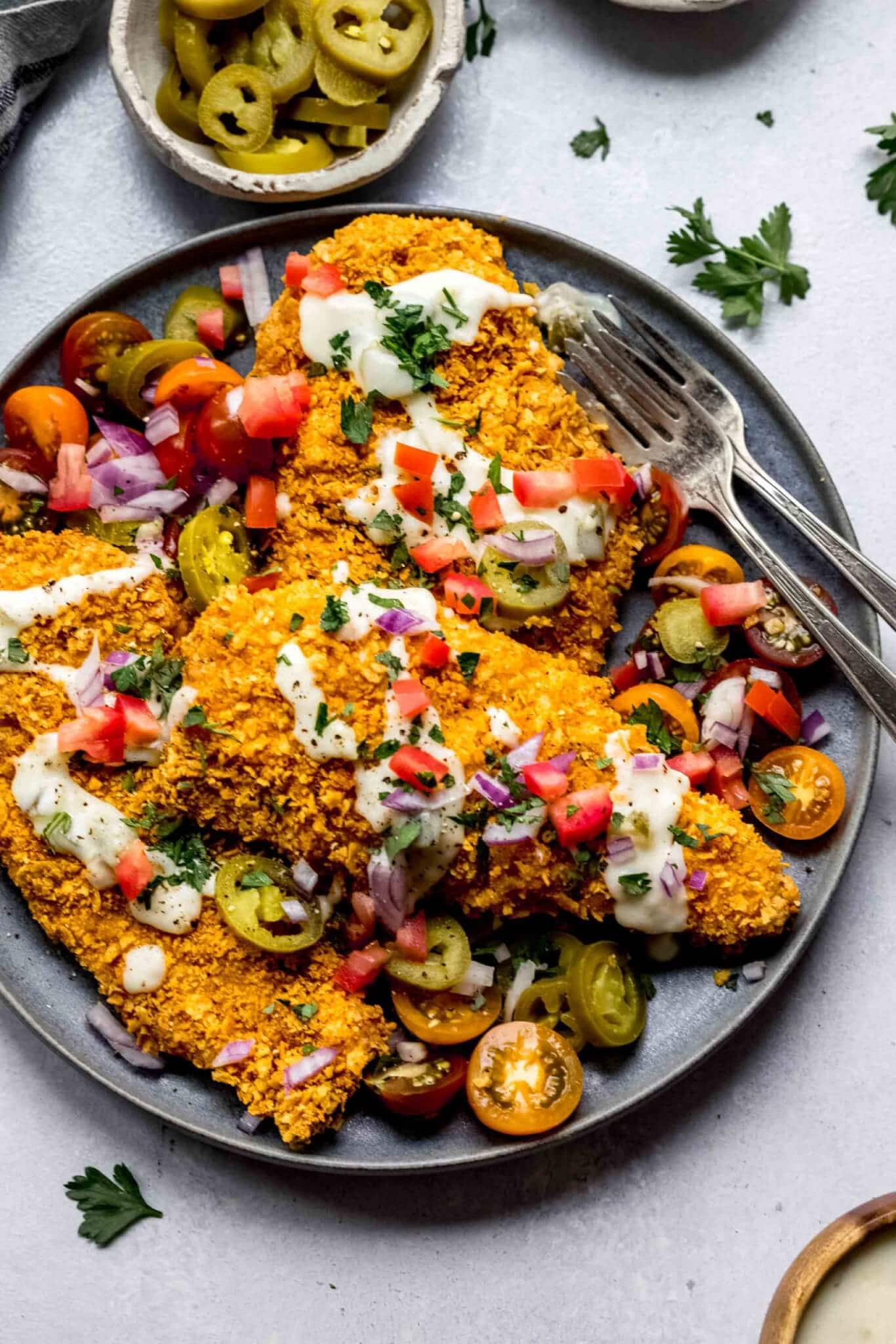Tortilla chip crusted chicken cutlets arranged on plate topped with queso, tomatoes and red onion.