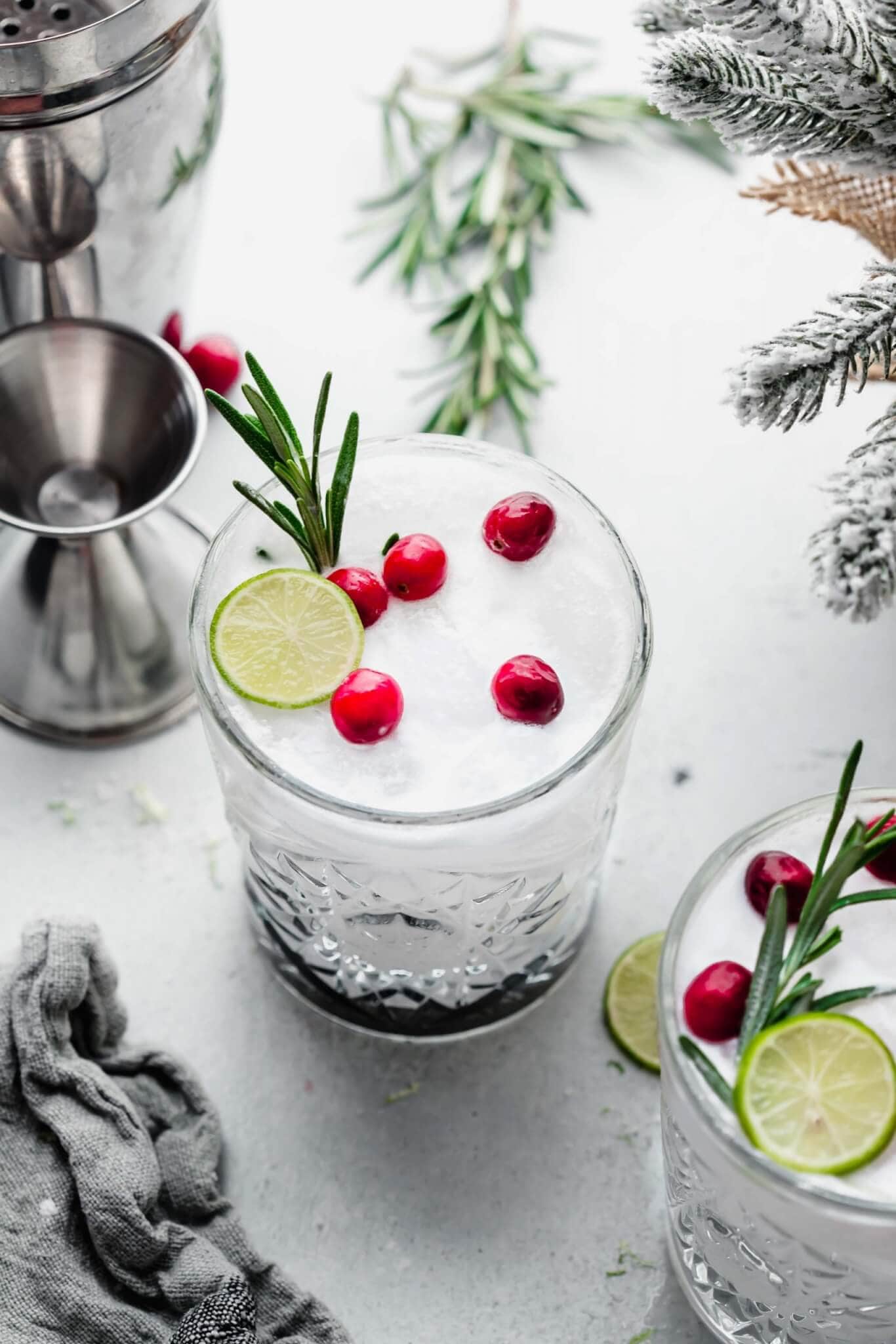 White Christmas Margarita topped with cranberries, lime slice, and rosemary next to jigger and towel