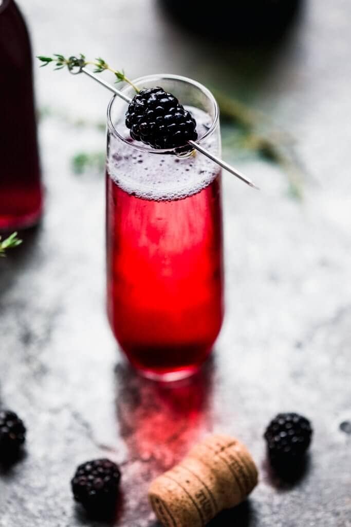 Shot of blackberry champagne cocktail garnished with blackberry and thyme sprig. 