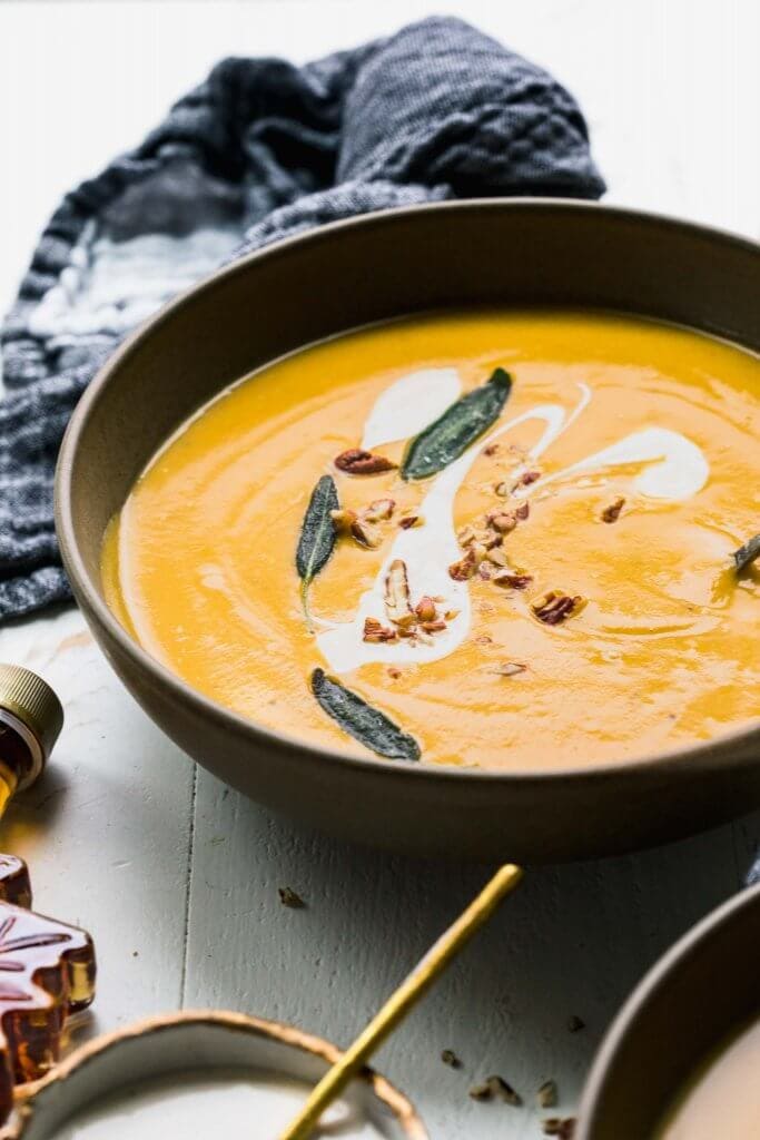 Butternut squash bisque in brown bowl topped with maple cream, walnuts and sage leaves.