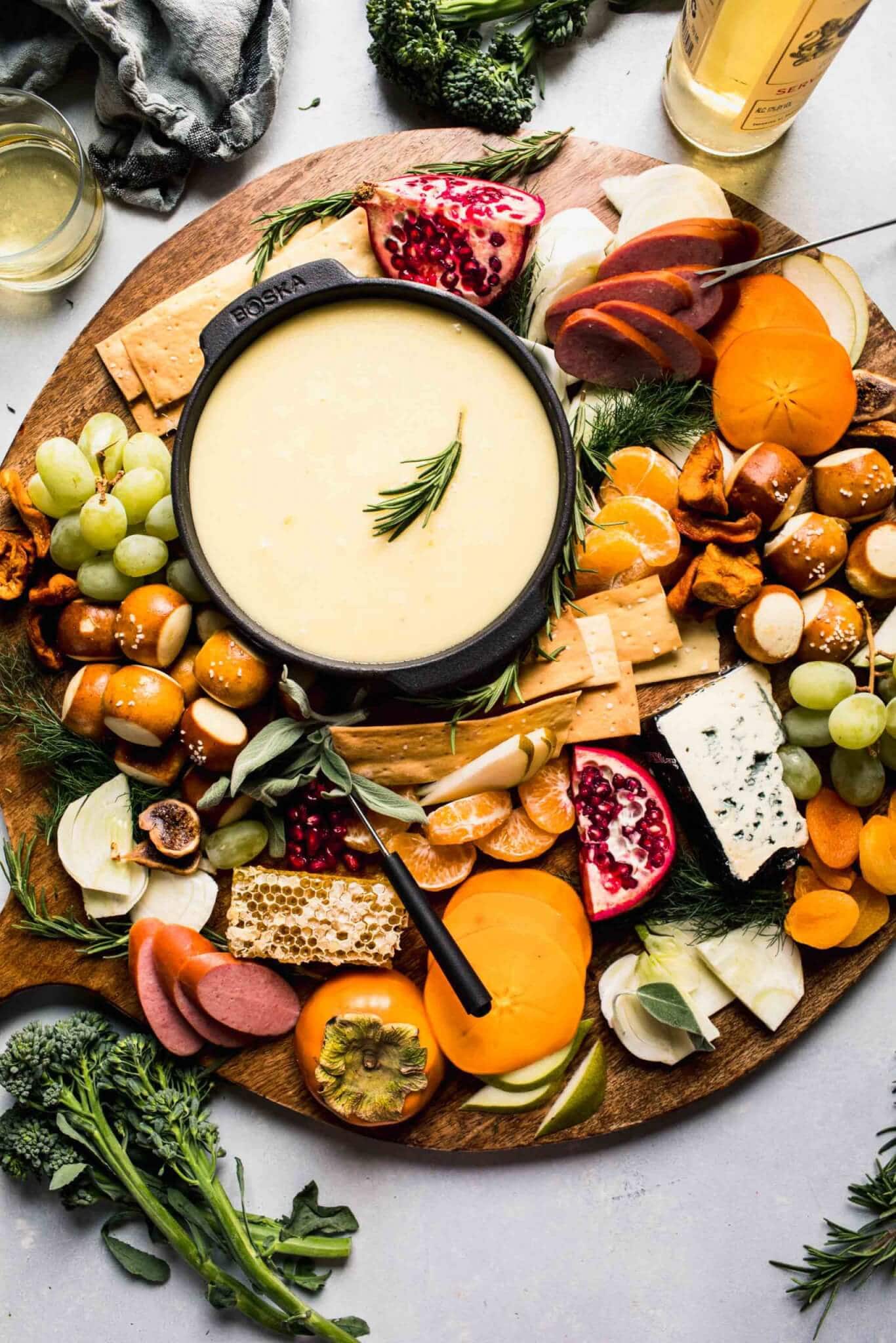 Fondue pot on cheese board surrounded by fruits, crackers and meats. 