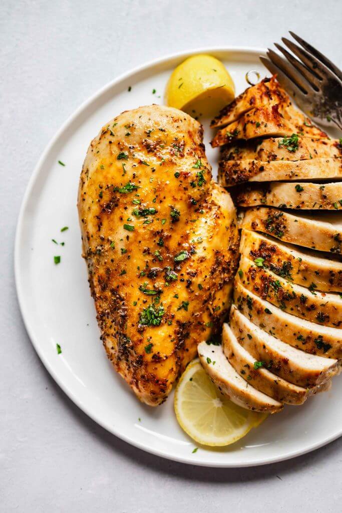 Overhead shot of baked chicken breast on white plate next to lemon slices and sliced chicken breast.