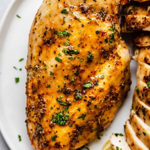 Overhead shot of baked chicken breast on white plate next to lemon slices and sliced chicken breast.