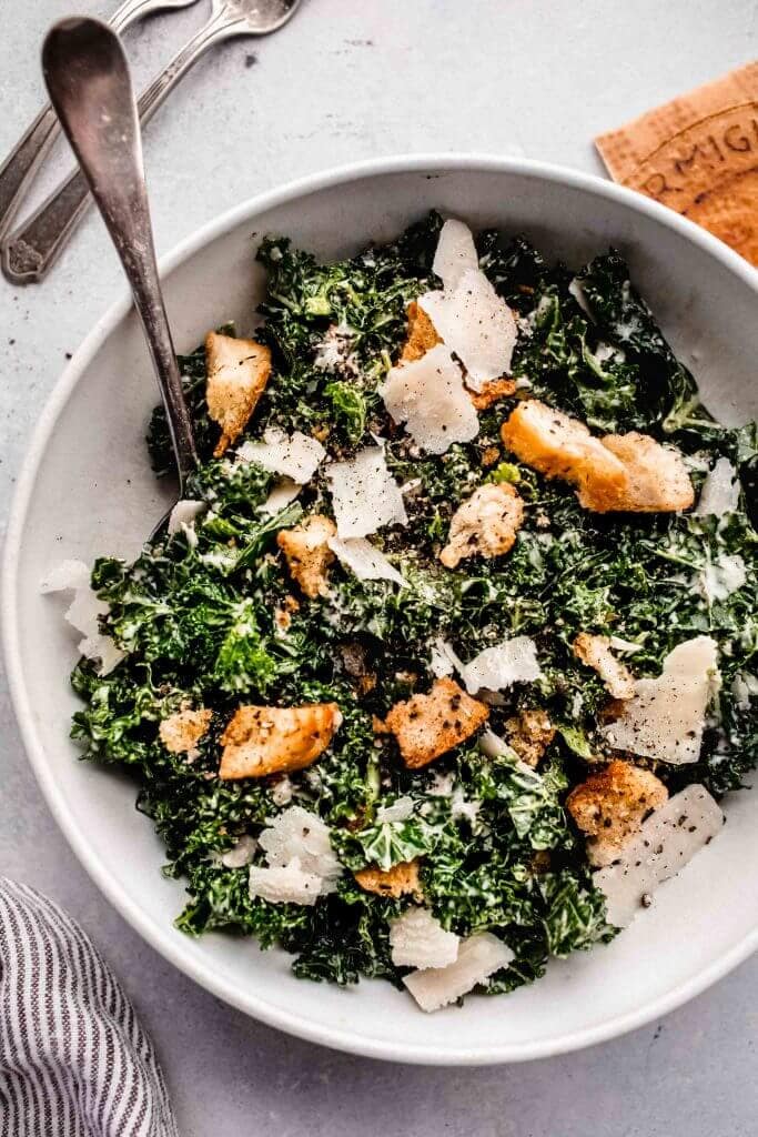 Overhead close up of bowl of kale caesar salad in white bowl with serving spoon.