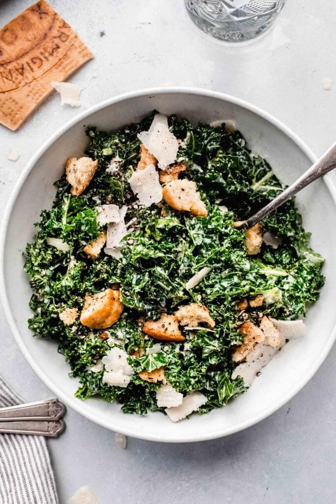 Overhead close up of bowl of kale caesar salad in white bowl with serving spoon.