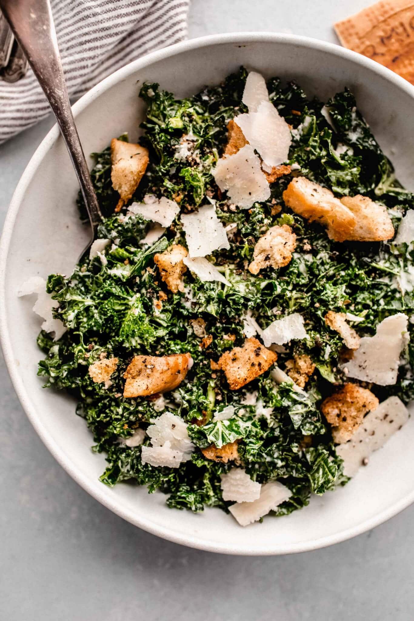 Overhead close up of bowl of kale caesar salad in white bowl with serving spoon.