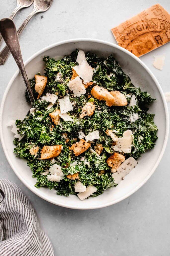 Overhead close up of bowl of kale caesar salad in white bowl with serving spoon.