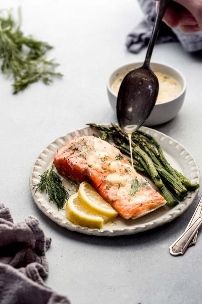 Top shot of spoon drizzling sauce onto salmon served with asparagus and lemon wedges on white table