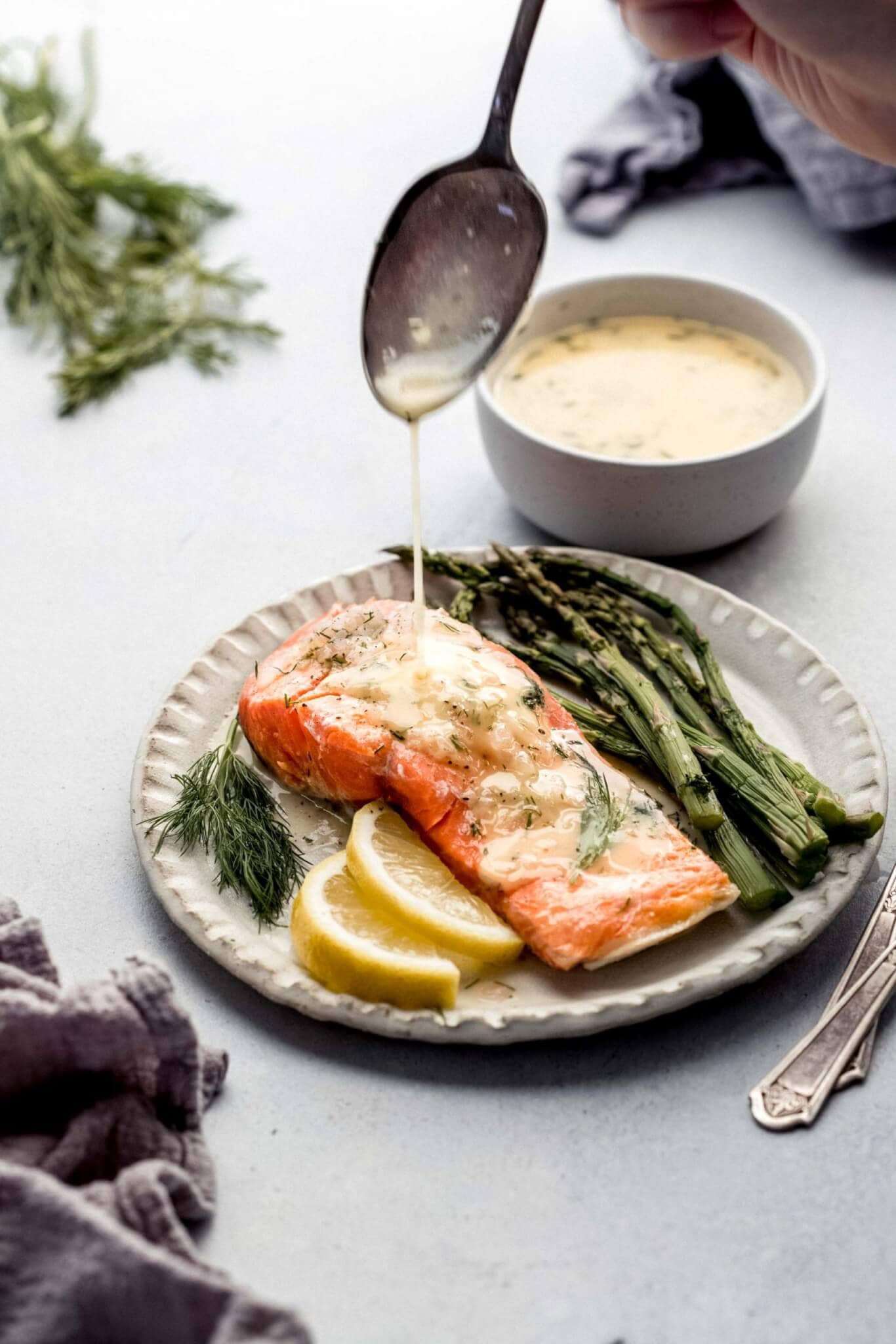 Top shot of spoon drizzling sauce onto salmon served with asparagus and lemon wedges on white table